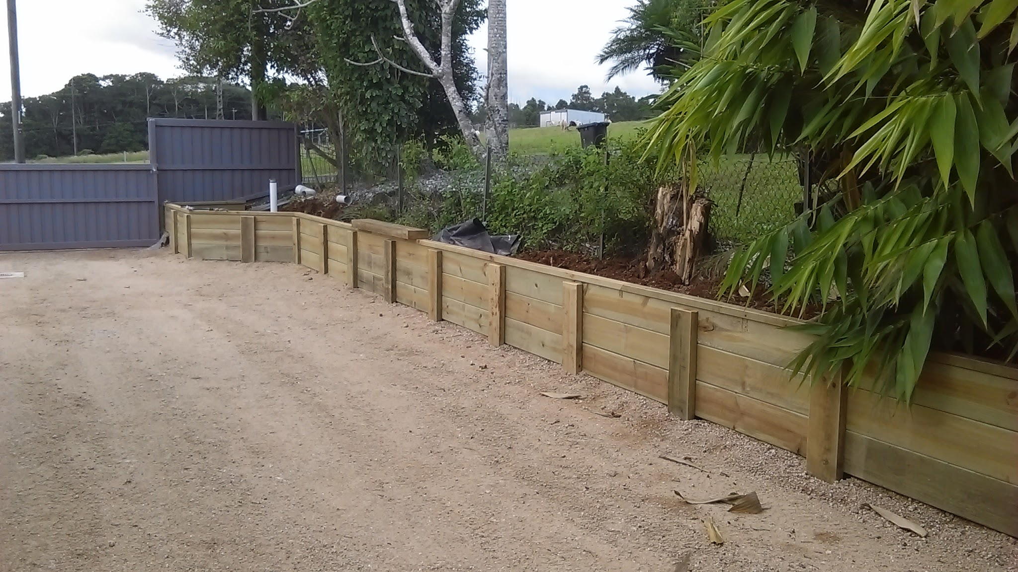 Treated timber sleeper retaining wall running along a gravel driveway, with a Colorbond fence and gate behind it