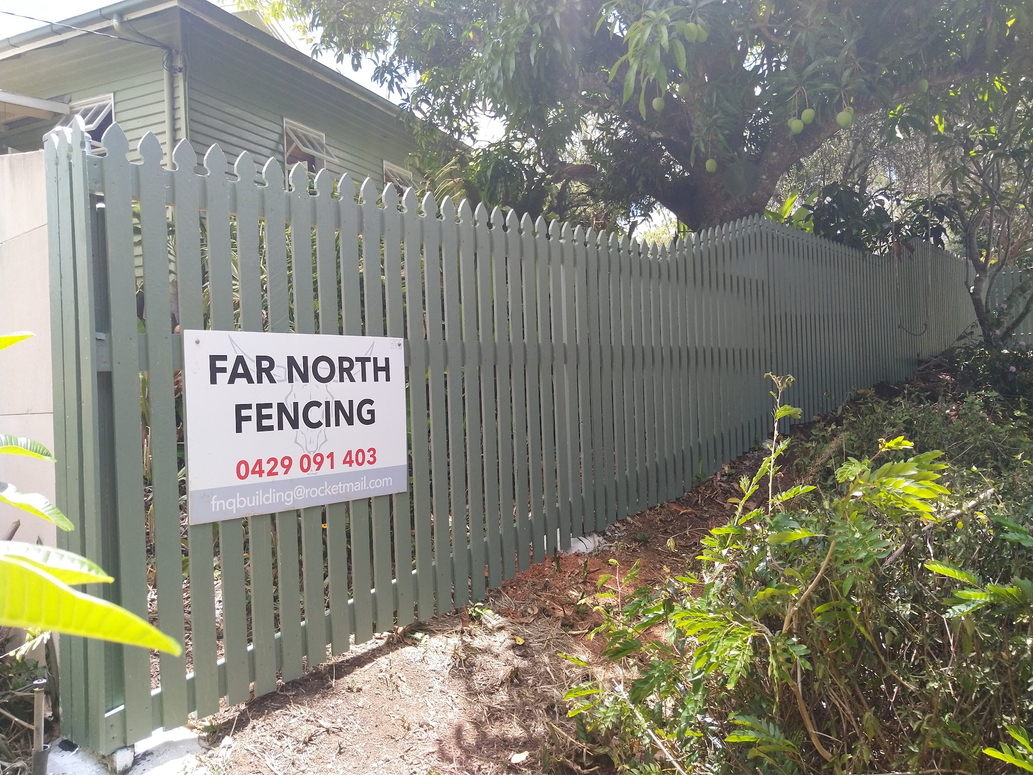 A sage green painted timber picket fence with the Far North Fencing job sign mounted on it