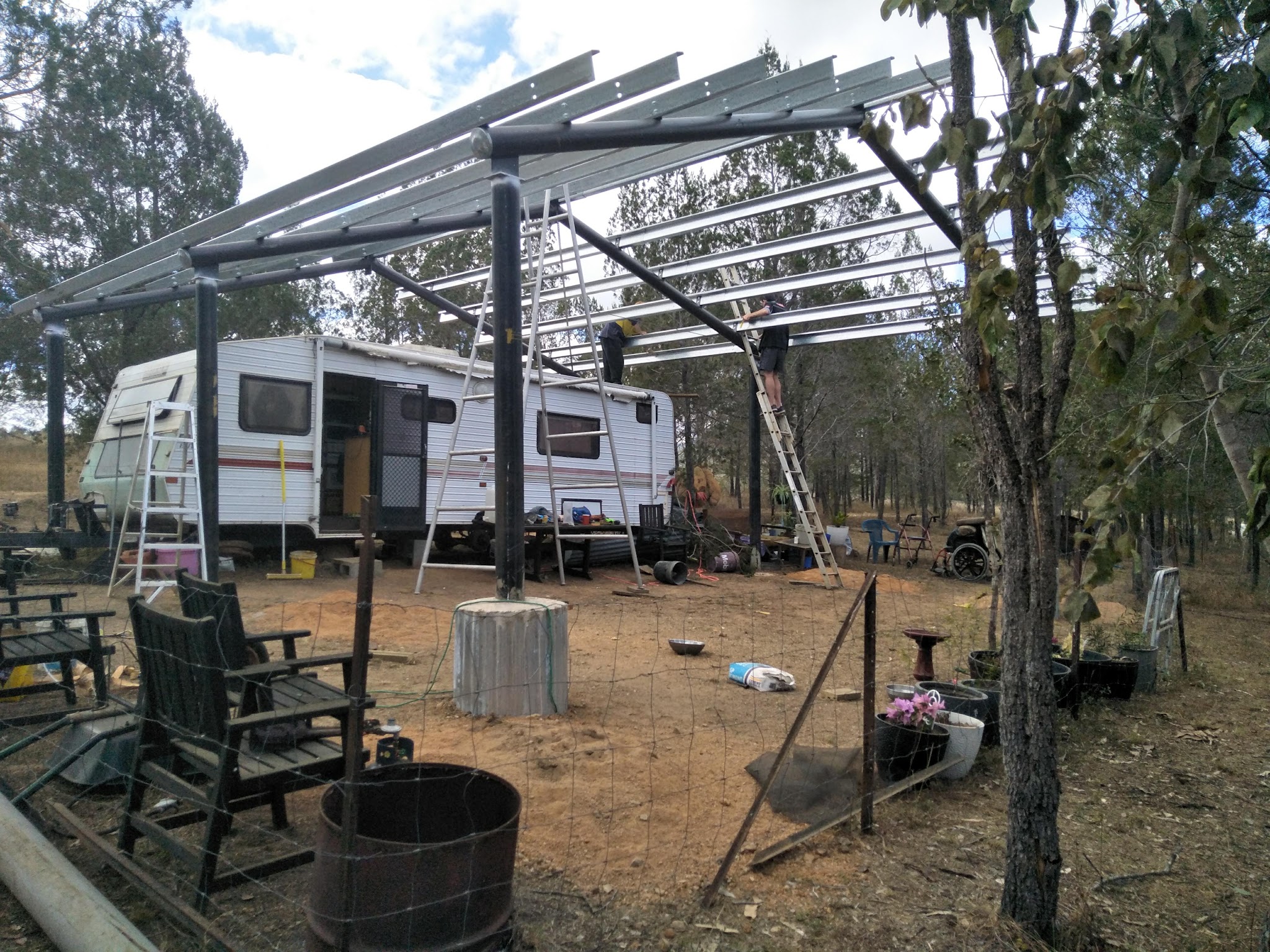 Steel carport frame being erected over a caravan, with two workers on ladders